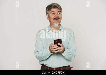 Mature man with grey beard smiling and using mobile phone isolated over white background Stock Photo