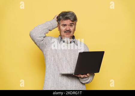 Mature man with grey beard frowning and using laptop isolated over yellow background Stock Photo
