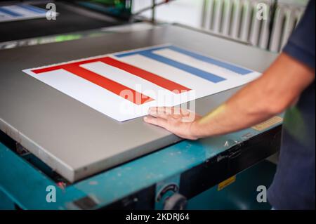 In the printing house, an experienced technician works on a screen printing machine. Production work. Check the print quality. Stock Photo