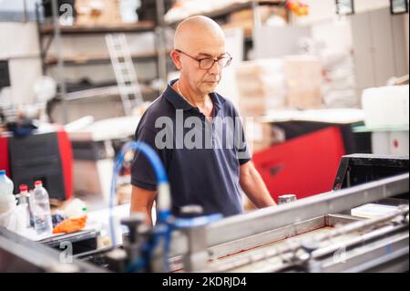 In the printing house, an experienced technician works on a screen printing machine. Production work. Check the print quality. Stock Photo