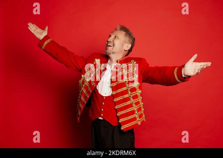 Portrait of mature man in costume of hussar posing isolated over red studio background. Artist Stock Photo