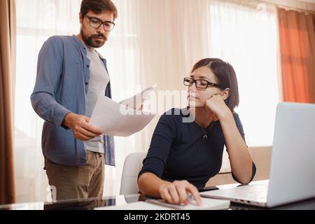 concept of home office, remote learning and online marketing. young couple using laptop and analyzing their finances with documents. Look at the papers. Stock Photo