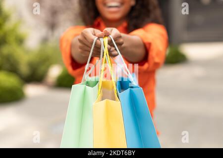Smiling millennial black woman in casual shows many of bags with order, enjoys great shopping in city, cropped Stock Photo