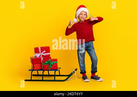 Full body photo of adorable small boy double thumb up christmas sleigh dressed stylish red knitted look isolated on yellow color background Stock Photo