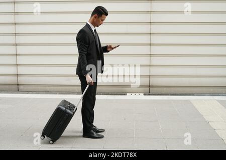 Elegant businessman checking e-mail on mobile phone while walking with suitcase Stock Photo