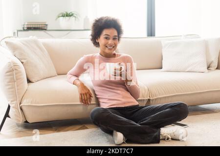 Smiling pretty relaxed millennial african american woman in casual enjoy cup of favorite hot drink, sit on floor Stock Photo
