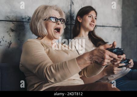 Two age generations women family, having fun enjoying play video game sitting on sofa. Woman playing video game with grandmother having fun on weekend Stock Photo