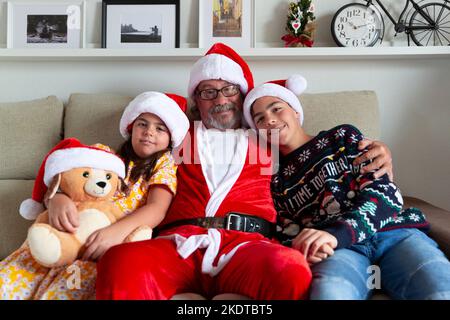 Christmas at home. Family portrait of grandfather dressed as Santa Claus hugging his grandchildren. Stock Photo