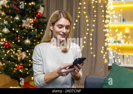 A young woman congratulates family and friends on Christmas holidays by phone. She is holding a mobile phone, typing a message, smiling, sitting alone at home on the sofa near the Christmas tree. Stock Photo