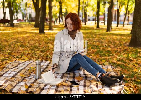 Concentrated smart caucasian pretty millennial lady student with red hair in raincoat sits on plaid, reads book Stock Photo