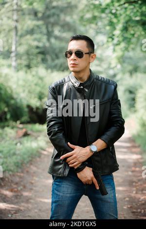 A man in dark glasses and a black leather jacket holds a gun in his hands Stock Photo