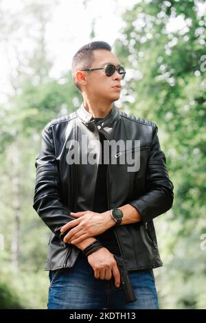 A man in dark glasses and a black leather jacket holds a gun in his hands Stock Photo