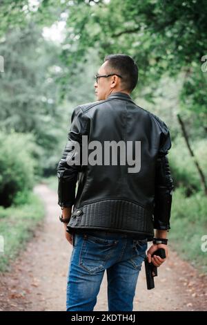 A man in dark glasses and a black leather jacket holds a gun in his hands Stock Photo