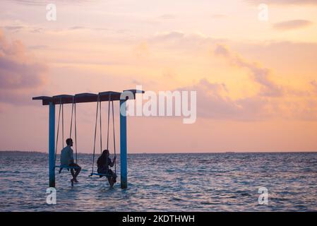 A picture of two couples sitting on a swing in the shallow sea of Maldives and looking at the beautiful sky during sunset Stock Photo