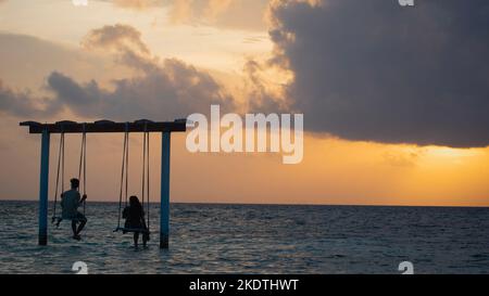 A picture of two couples sitting on a swing in the shallow sea of maldives and looking at the beautiful orange sky during sunset Stock Photo