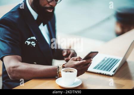 View of a bearded black man entrepreneur in a street cafe, with a selective focus on his hand typing a message on the smartphone; a cup of espresso co Stock Photo