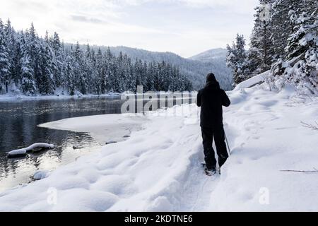 Man Snowshoeing on Trail Along Partly Frozen Lake in Winter Stock Photo