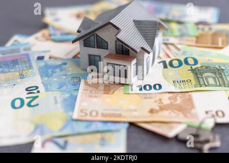 Small toy house is lies on a set of green monetary denominations of 100 euros. A lot of money forms an infinite heap. Stock Photo