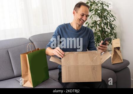 Present from distant friend. Happy millennial man unpack open box with birthday gift surprise received by mail. Smiling young guy has pleasure to Stock Photo