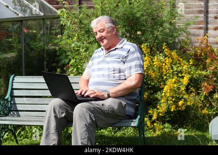 Senior or elderly man sitting outside in his garden with his laptop computer on his lap. Happy working in the sunshine. Stock Photo