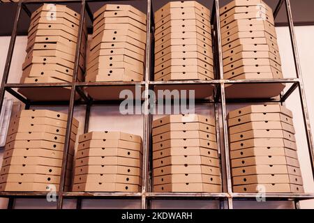 food delivery service. tall stacks of flat brown cardboard pizza boxes on metal shelf ready for delivery. craft packing boxes with pizza in stock in s Stock Photo
