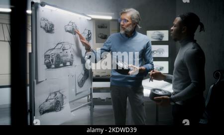 Two diverse automotive male designers holding small wheels, developing futuristic electric car design, standing near whiteboard with sketches. They working in car design development modern studio. Stock Photo