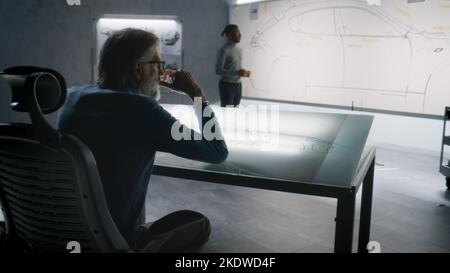 Two diverse automotive male engineers developing sketch of futuristic electric car design. One drawing with marker on whiteboard, other working at the light table. Car design development studio. Stock Photo