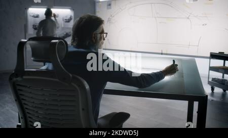 Two diverse automotive male engineers developing sketch of futuristic electric car design. One drawing with marker on whiteboard, other working at the light table. Car design development studio. Stock Photo
