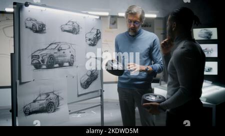 Two diverse automotive male designers holding small wheels, developing futuristic electric car design, standing near whiteboard with sketches. They working in car design development modern studio. Stock Photo
