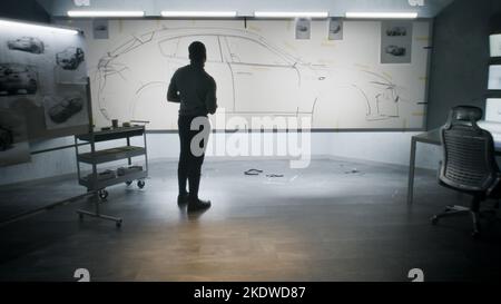 Back view of automotive african american male designer drawing sketch blueprint with marker, developing futuristic electric car design on whiteboard. Work in modern car design development studio. Stock Photo