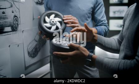 Two diverse automotive male designers holding small wheels, developing futuristic electric car design, standing near whiteboard with sketches. They working in car design development modern studio. Stock Photo