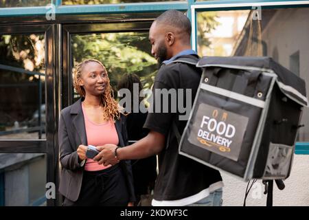 Woman paying for food order with credit card pos terminal, contactless payment. African american courier delivering restaurant takeaway meal, smiling company employee receiving lunch Stock Photo