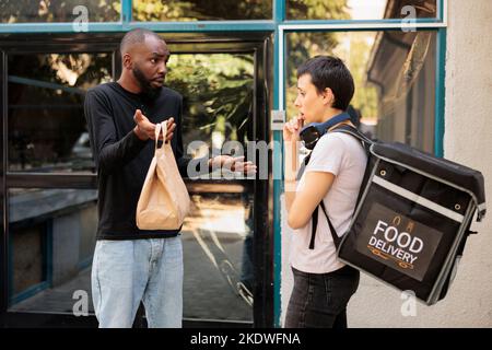 Food delivery bad service, unsatisfied client holding order package, courier and angry customer. Woman giving unhappy employee takeaway meal paper bag near office building outdoors Stock Photo
