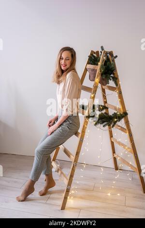 Portrait of modern middle aged woman at wooden ladder, decorated with fir branches, garland for Christmas. Place for text, light wall. Scandinavian st Stock Photo