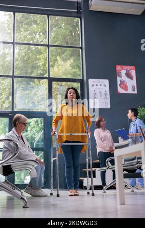 Senior physician attending physiotherapy with injured patient, doing rehabilitation examination and trying to walk with walking frame. Asian woman with leg fracture working on physical recovery. Stock Photo