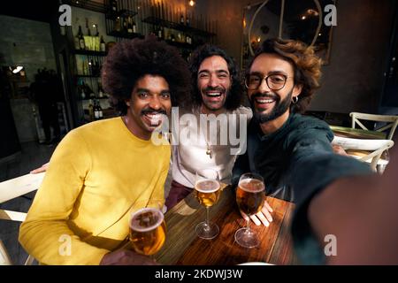 Horizontal selfie of three male friends toasting happily in a bar, looking at camera with big smiles. Stock Photo
