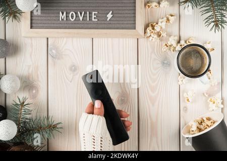 Cropped woman hands in cozy sweater holding remote control, nameplate movie, cup of coffee, bucket of popcorn on decorative xmas wooden background Stock Photo