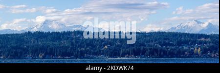 A panoramic view across the water of snow capped mountains on the Olympic Peninsula in Washington with clouds in the sky. Stock Photo