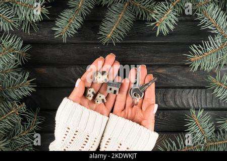 Cropped woman hands in white sweater holding metallic keys and miniature house model toys in evergreen twigs decoration on wooden view. Housewarming Stock Photo