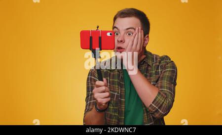 Wow reaction. Impressed man blogger taking selfie on mobile phone selfie stick, communicating video call online with subscribers. Young teen guy boy 20s isolated alone on yellow studio wall background Stock Photo