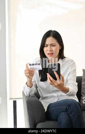 Unhappy and dissatisfied Asian woman sitting on sofa, looking at her smartphone screen, having problem with mobile banking application. female with ph Stock Photo