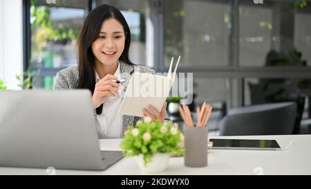 Charming young Asian businesswoman checking some details on her notebook, working in her office. Stock Photo