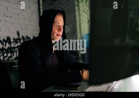 Close-up of wanted hackers coding virus ransomware using laptops and computers sitting at desk in dark room. Hooded hacker typing on laptop keyboard. Stock Photo