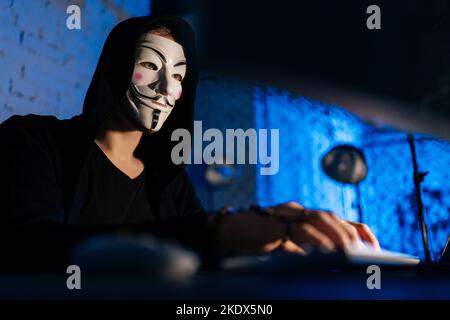 Low-angle view of unrecognizable wanted hacker male wearing anonymous mask typing on wireless keyboard breaking password using laptop and desktop pc. Stock Photo