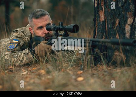 Ukrainian soldier dressed military uniform lying on ground with sniper rifle with silencer at front line. Handsome brave confident man at war Stock Photo
