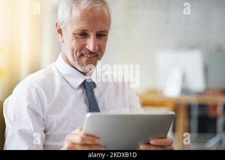 Keeping track of his online interests. a smiling mature businessman using a digital tablet while standing in an office. Stock Photo