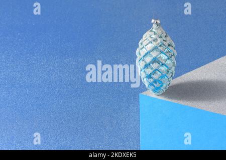 Optical illusion of a vintage christmas bauble in the shape of a pine cone standing in balance on a paper cube against a blue background. Stock Photo