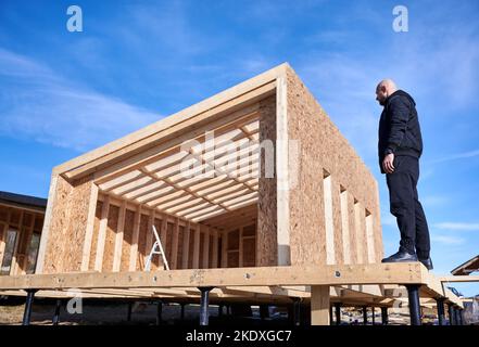 Male designer building wooden frame house on pile foundation. Bald man standing on construction site, inspecting quality of work on sunny day with blue sky on background. Stock Photo