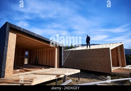 Inspector building wooden frame house on pile foundation in Scandinavian style barnhouse. Bald man standing on construction site, inspecting quality of work on sunny day with blue sky on background. Stock Photo