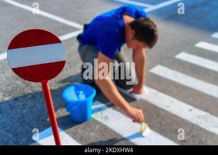 Road service worker paints striped pedestrian crossing on asphalt with paint brush. Markings for pedestrians on roadway. Safe transition for people. Real workflow.. Stock Photo